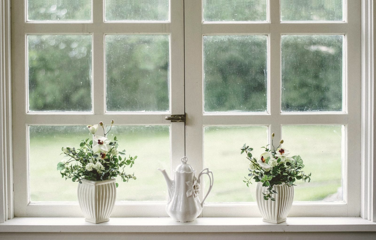 Vita blomsterarrangemang och en kaffekanna i porslin på en fönsterbräda i mjukt dagsljus